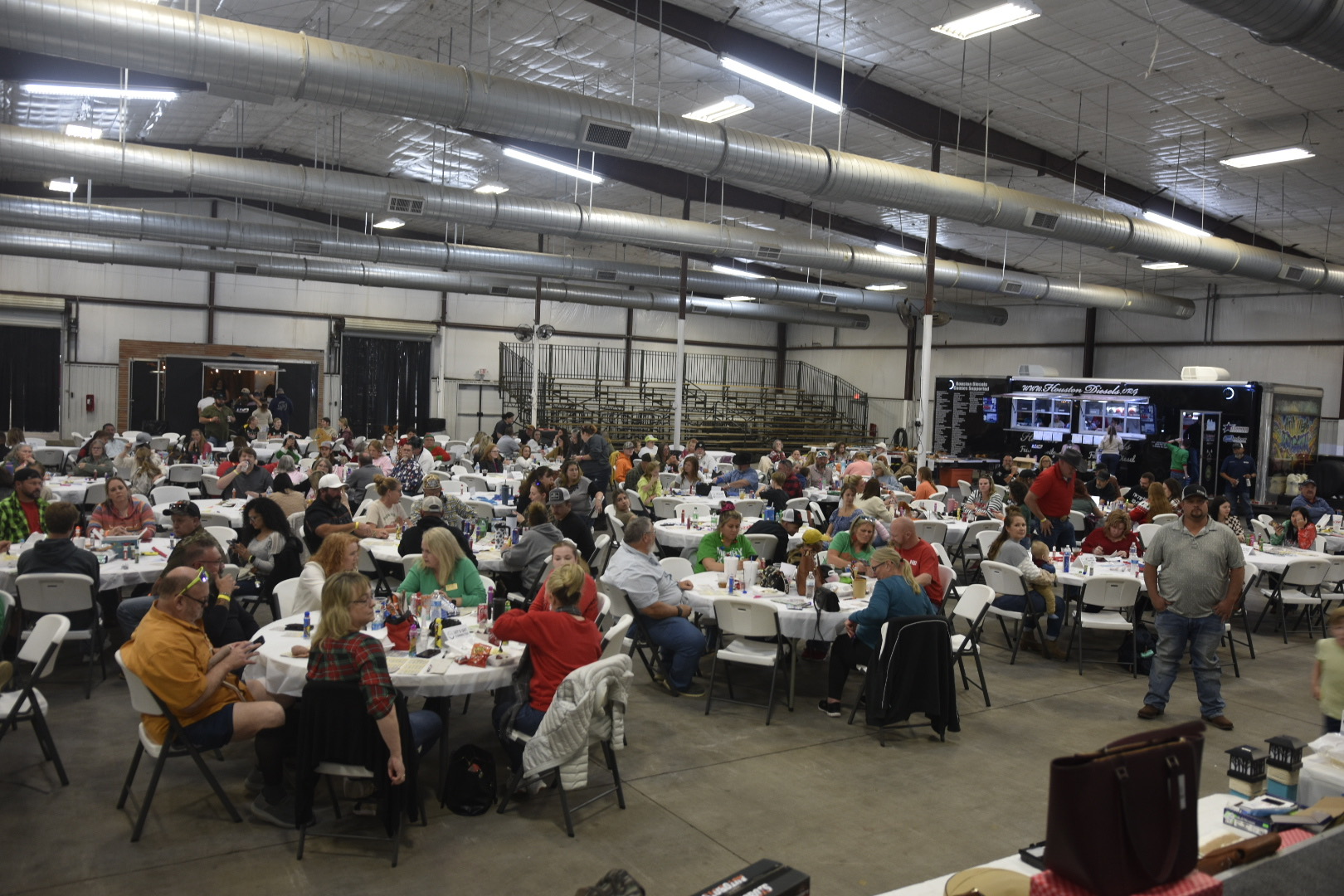 Large indoor event with many people seated at tables, eating and socializing, in a spacious hall with industrial ceiling and lighting.