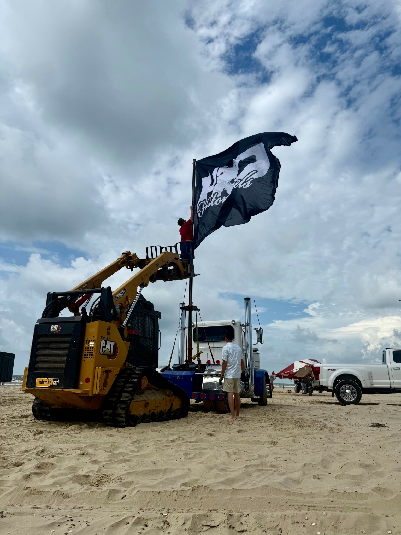 Construction equipment and trucks on a sandy area under a cloudy sky, with a person holding a flag.