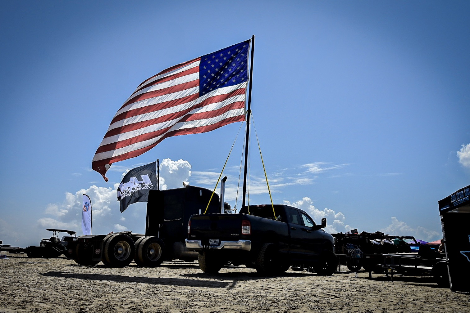 Black truck with American flag on a pole, parked on a dirt surface under a blue sky with clouds.