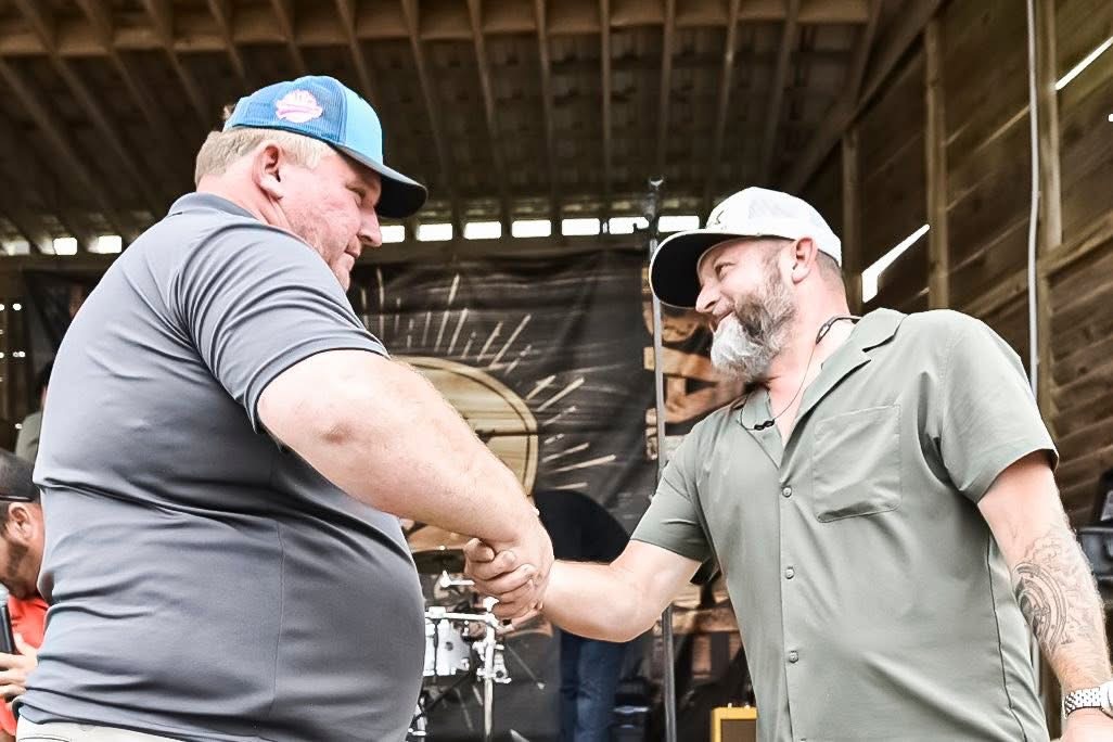 Two men shaking hands inside a wooden structure, one wearing a blue cap and gray shirt, the other a white cap and green shirt.