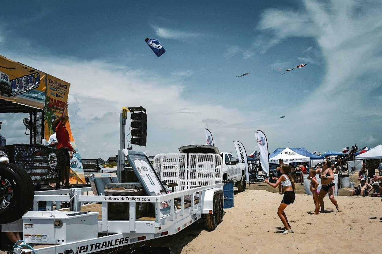 Beach scene with people, tents, and vehicles under a partly cloudy sky, with kites flying overhead.