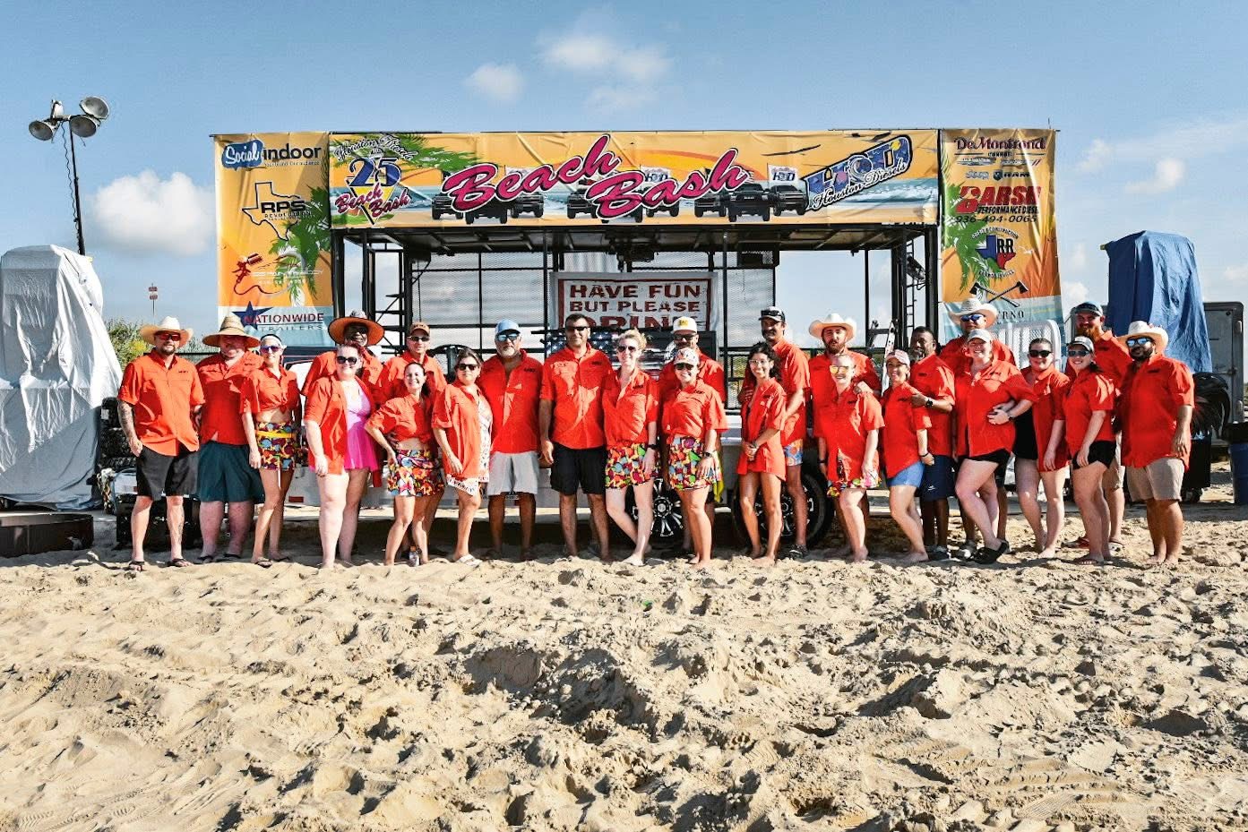 Group of people in red shirts standing on sandy beach in front of a colorful stage with banners, under a clear blue sky.