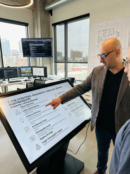 Man in glasses and blazer points at a large touchscreen display in a modern office with city view windows.