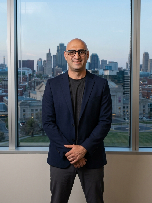 Man with glasses in a dark blazer and black shirt standing in front of a large window with cityscape view.
