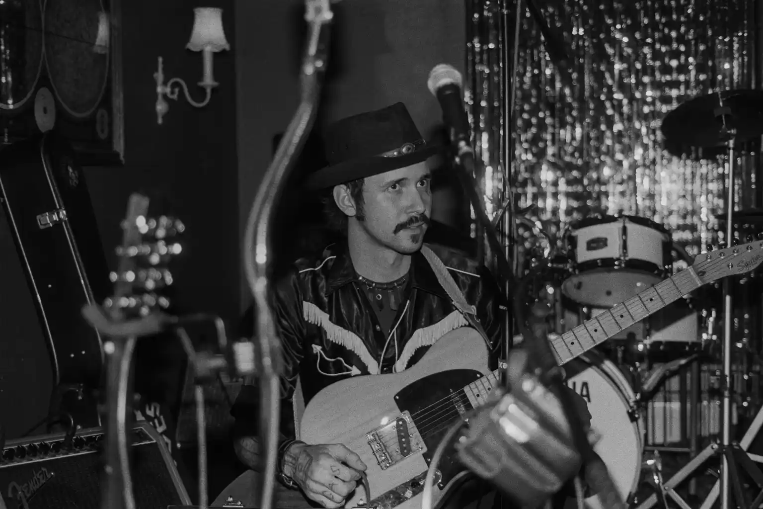 Man with a mustache wearing a hat and leather jacket playing guitar on stage with musical instruments and curtains in background.