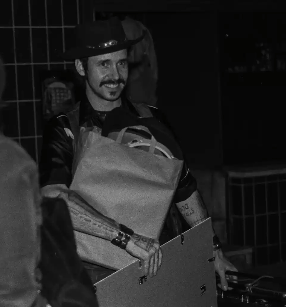 Man with a beard and mustache wearing a hat, smiling, holding a large bag, in a dark indoor setting.