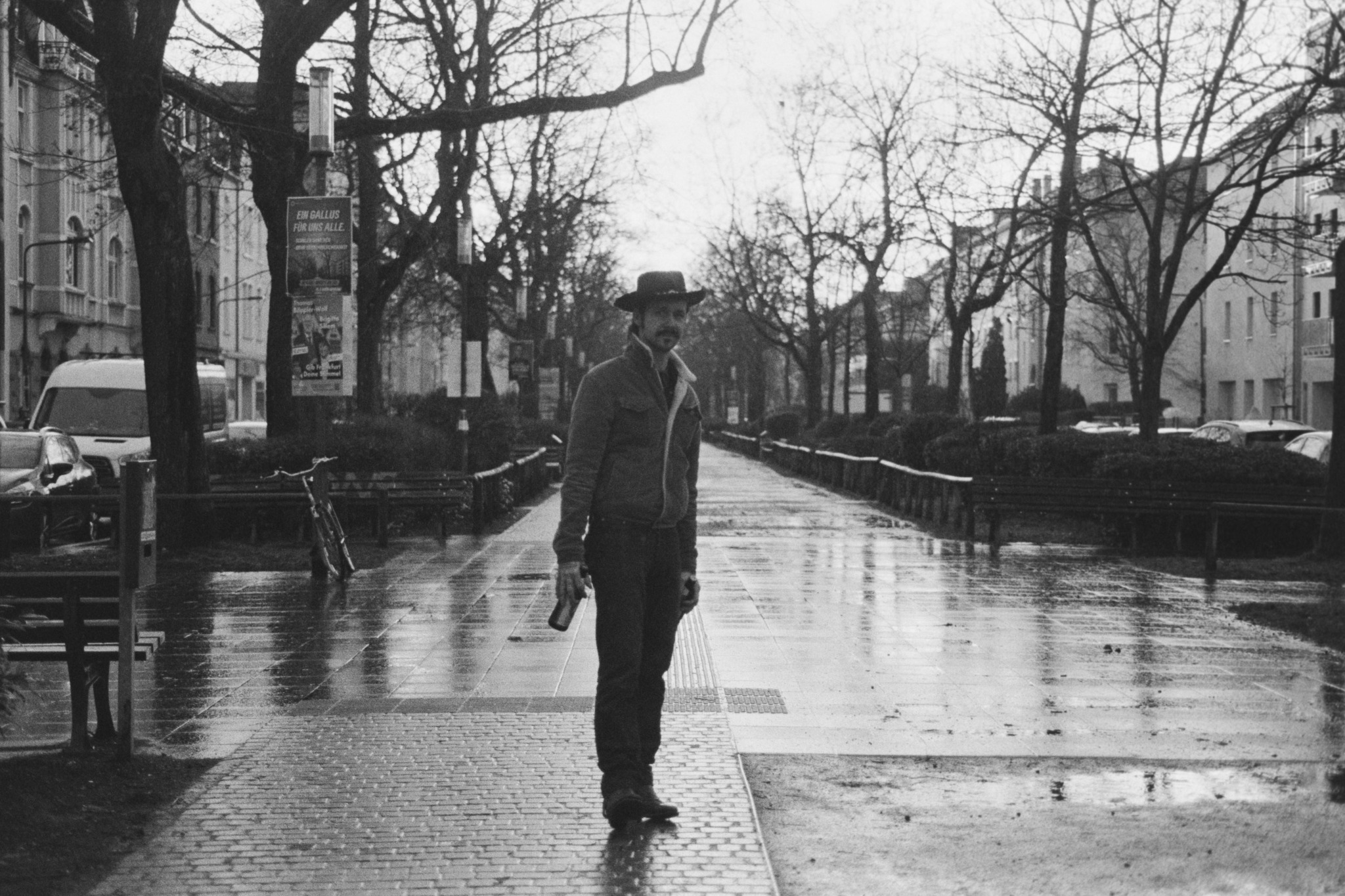 Man standing on wet sidewalk in park with trees and buildings in background, black and white photo.