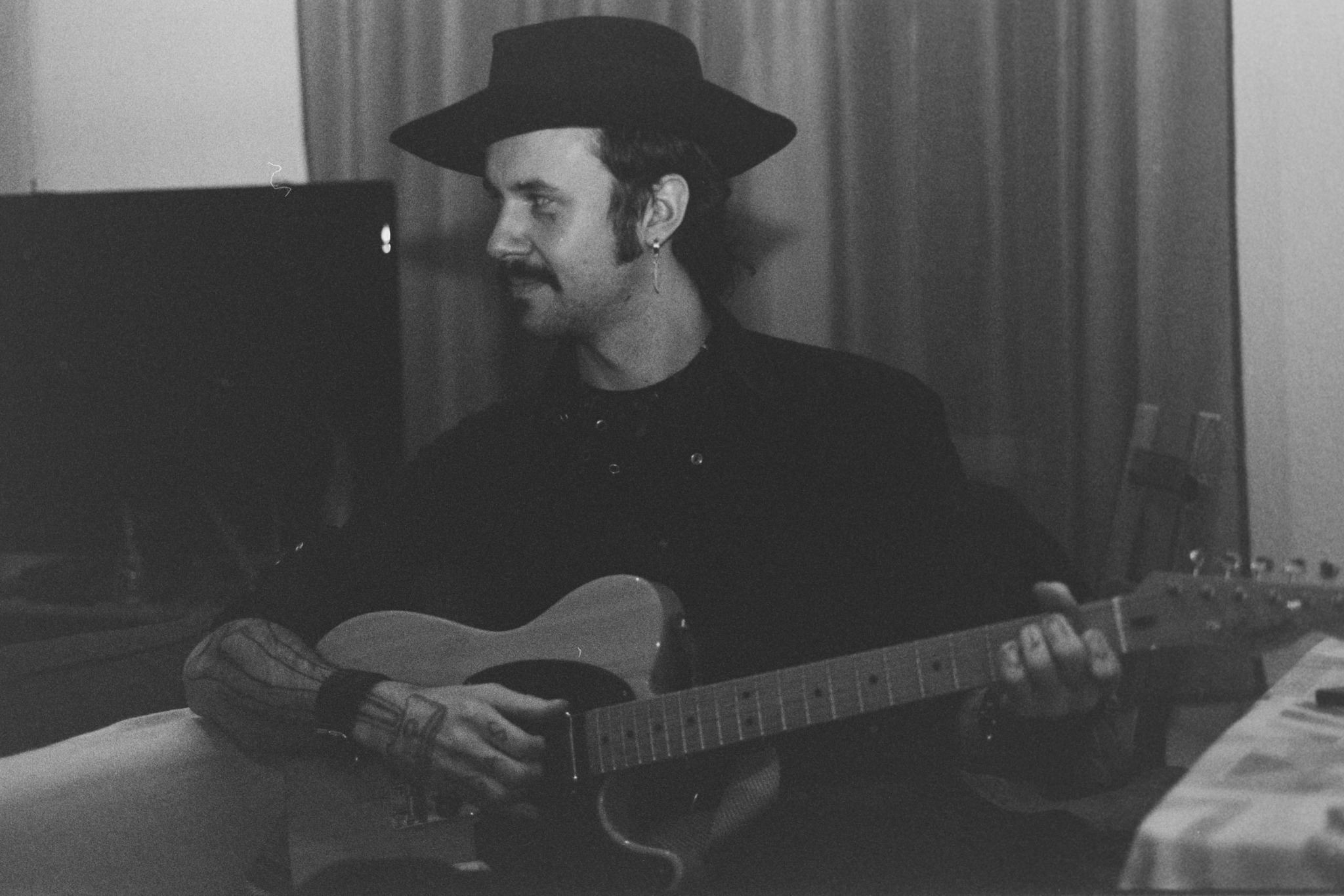 Person wearing a hat playing an acoustic guitar, sitting indoors with curtains in the background.