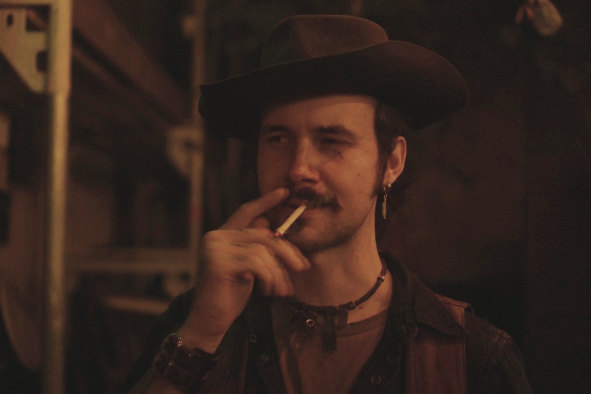 Man with a hat and earring smokes a cigarette in a dimly lit setting.