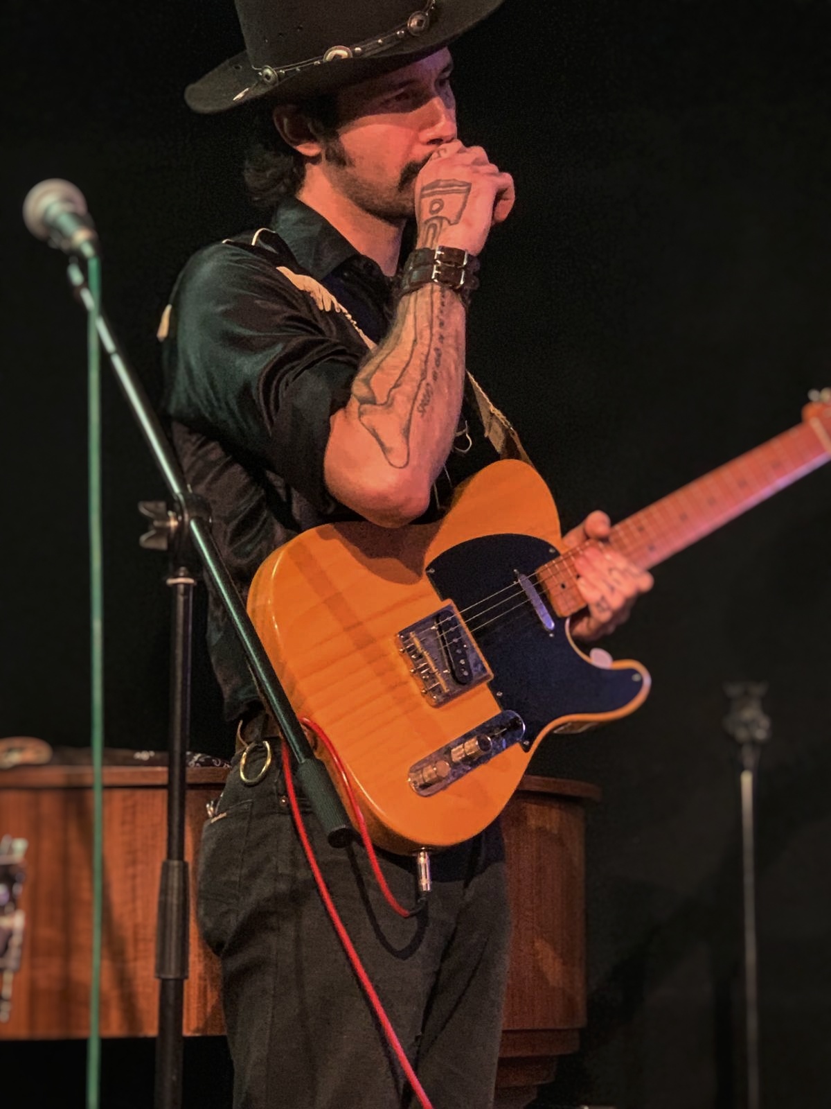 Man with a hat playing an electric guitar on stage, holding a microphone, with a dark background.
