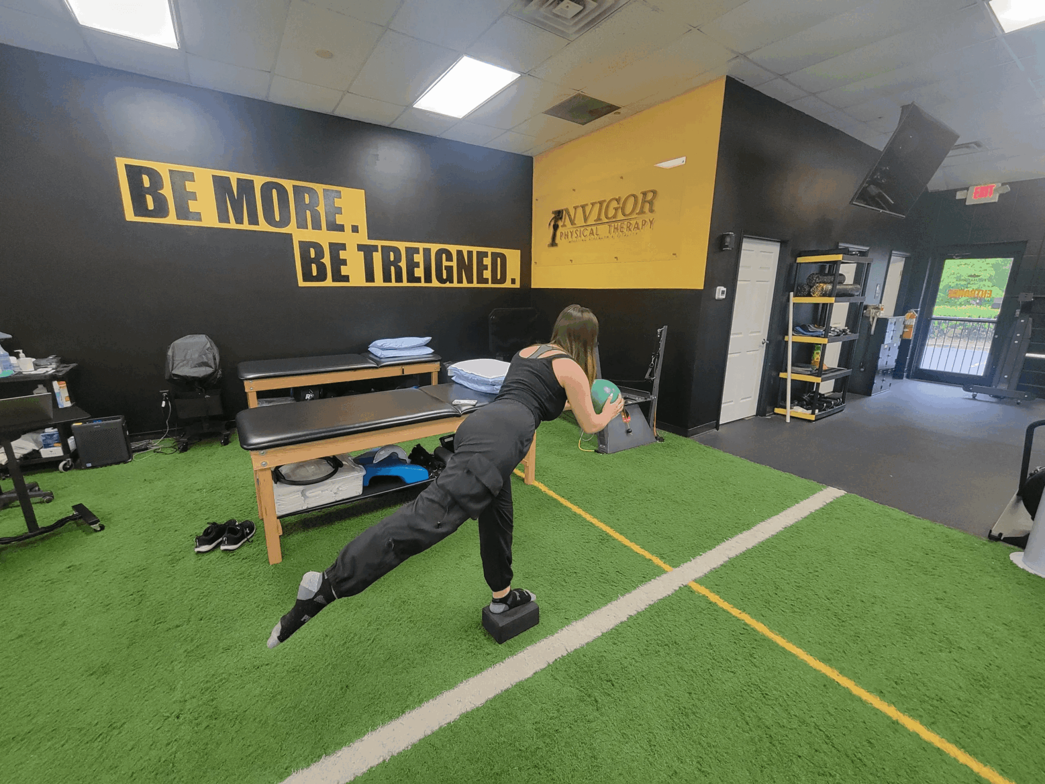 Young person in black workout clothes pushes a weighted sled on green turf inside a gym. Motivational signs are on the wall.