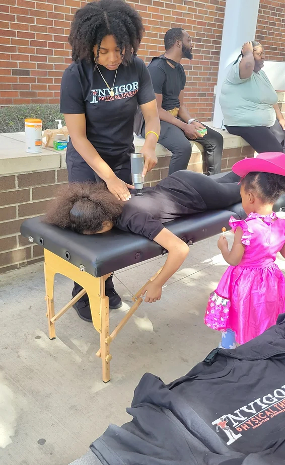 Person receiving a massage outdoors on a black massage table, with children and adults nearby, brick wall in background.