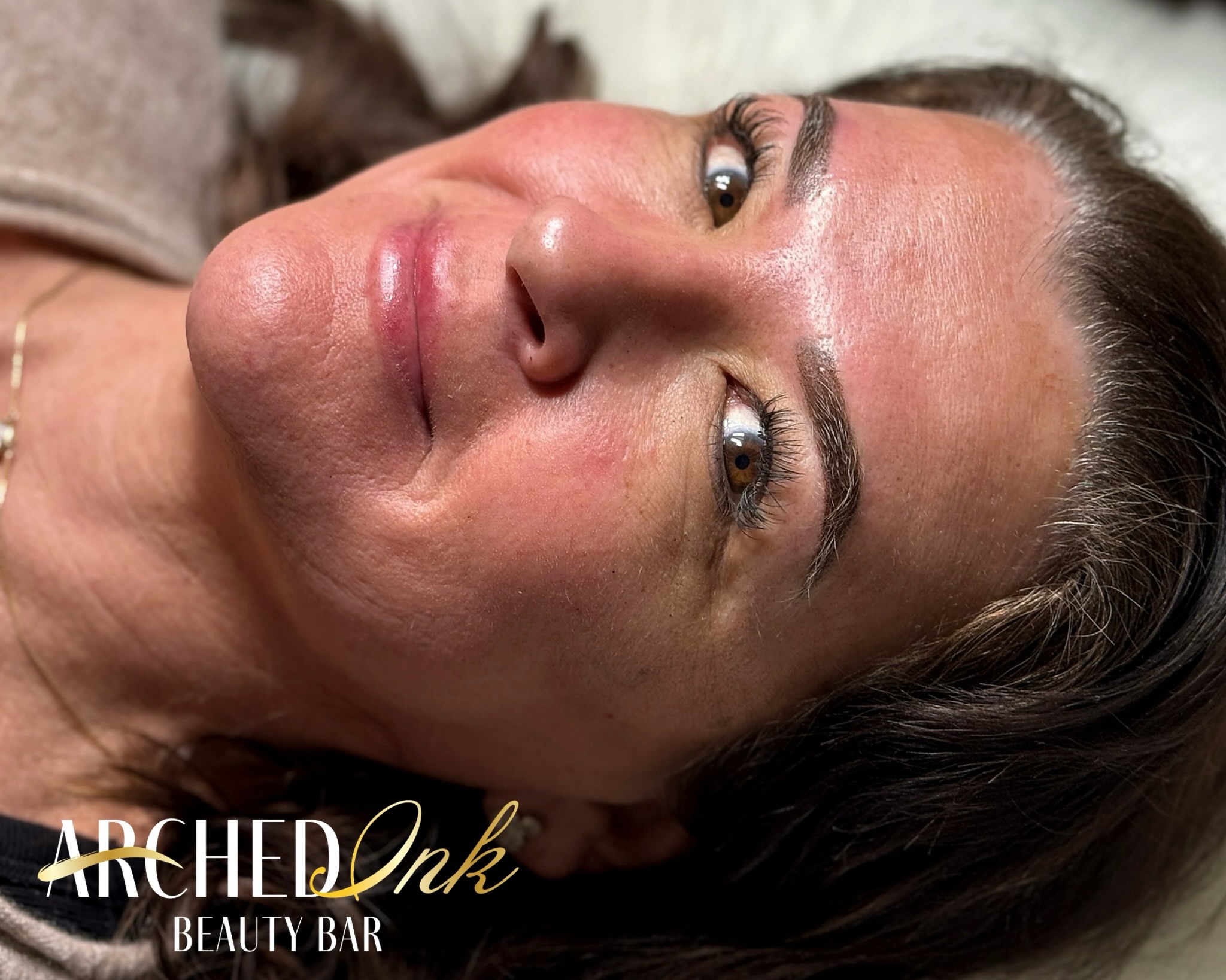 Close-up of a woman's face with makeup, lying down, with text 'Arched Ink Beauty Bar' at the bottom.