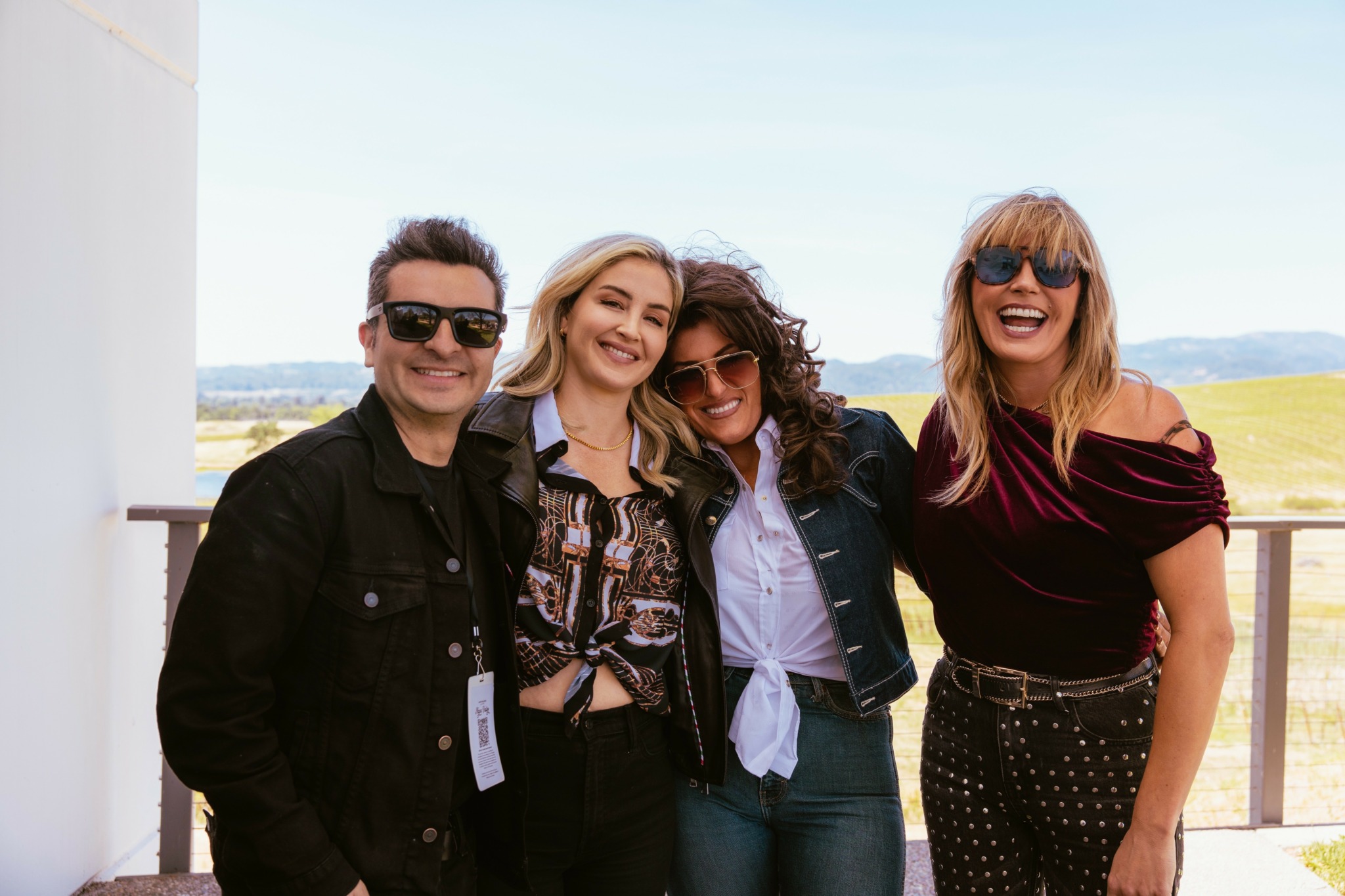 Four people smiling and standing close together outdoors on a balcony with a landscape background, sunny weather.