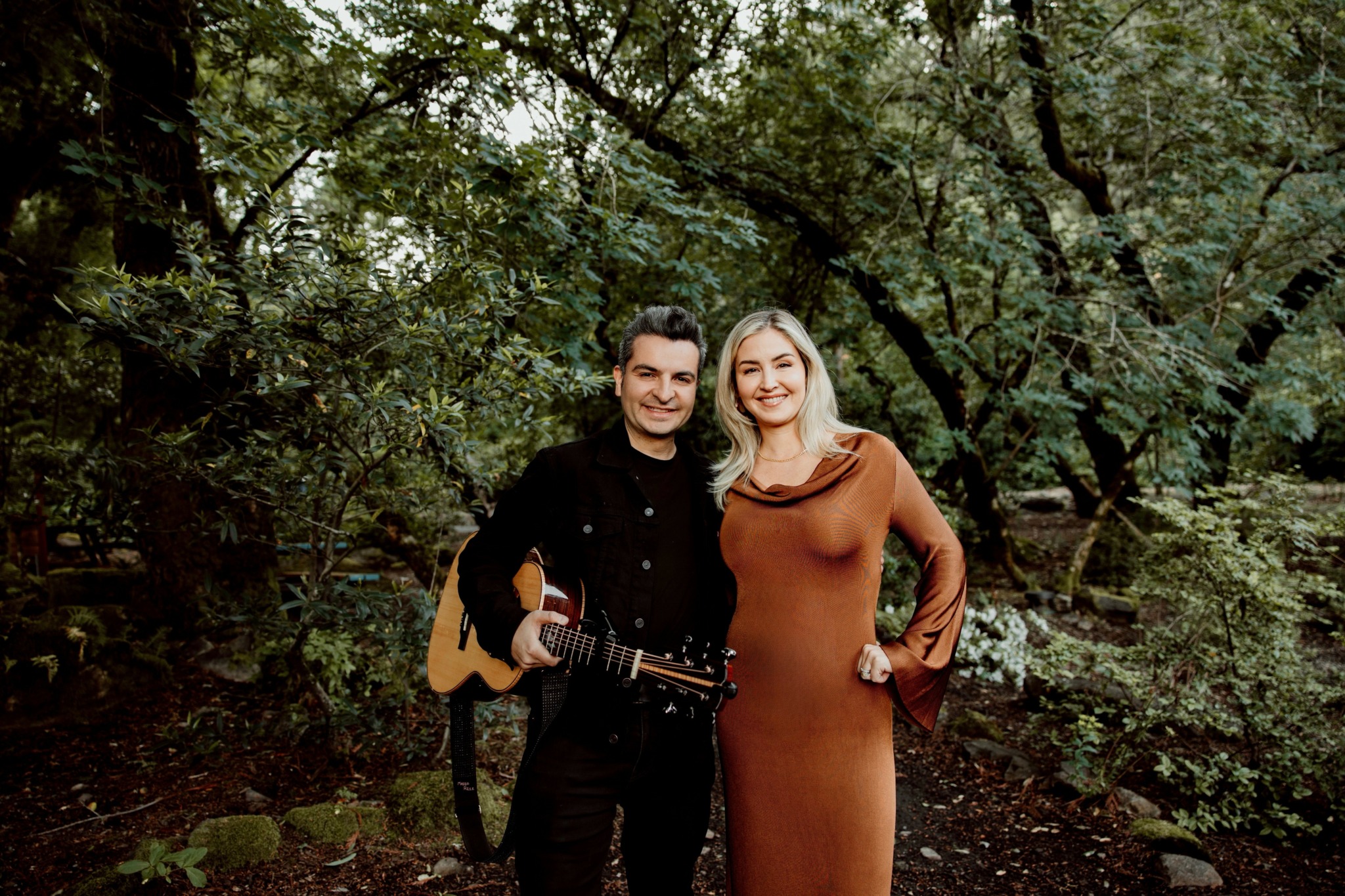 Two people standing outdoors among trees, smiling, with one holding a guitar, in a natural setting.