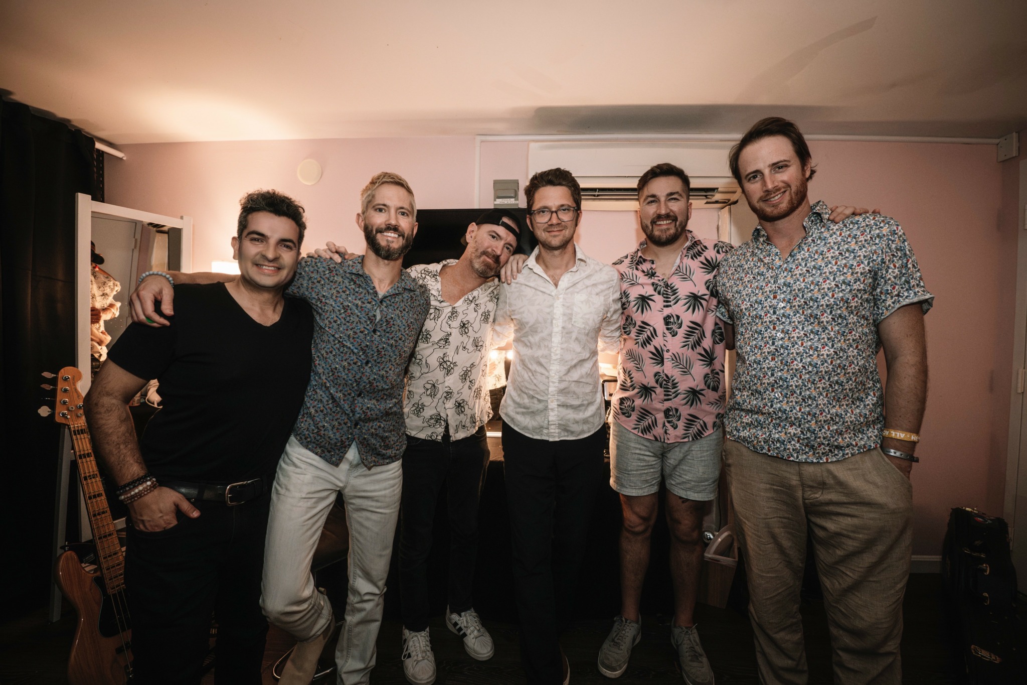 Group of seven men standing indoors, smiling, with a pink wall and window behind them.