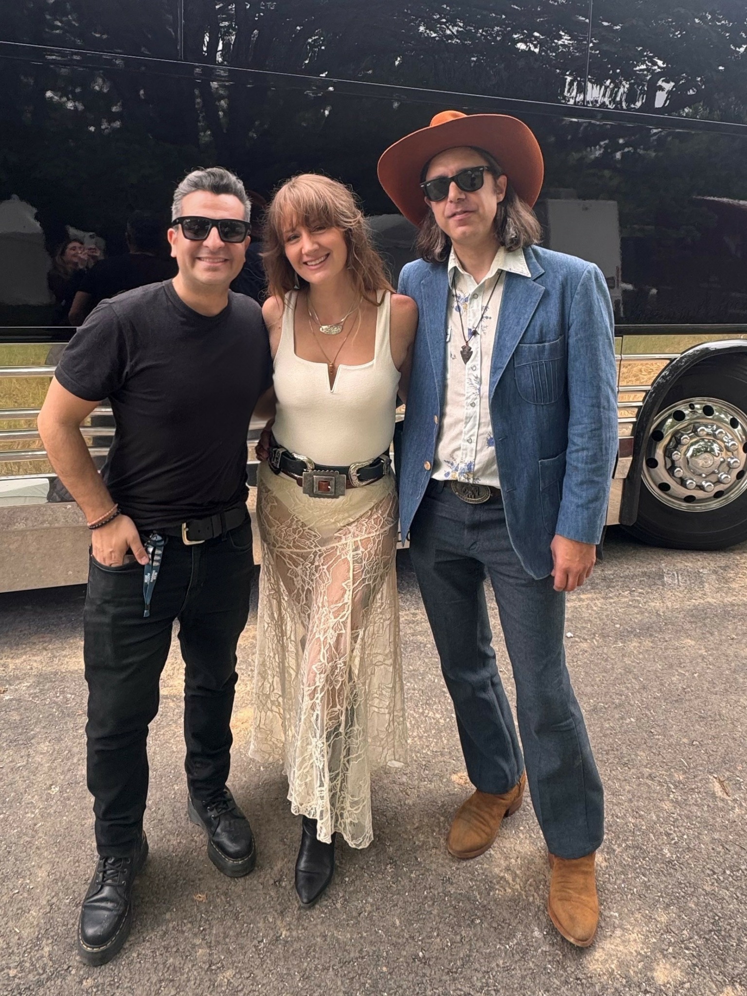 Three people standing outdoors, smiling, with a vehicle in the background.