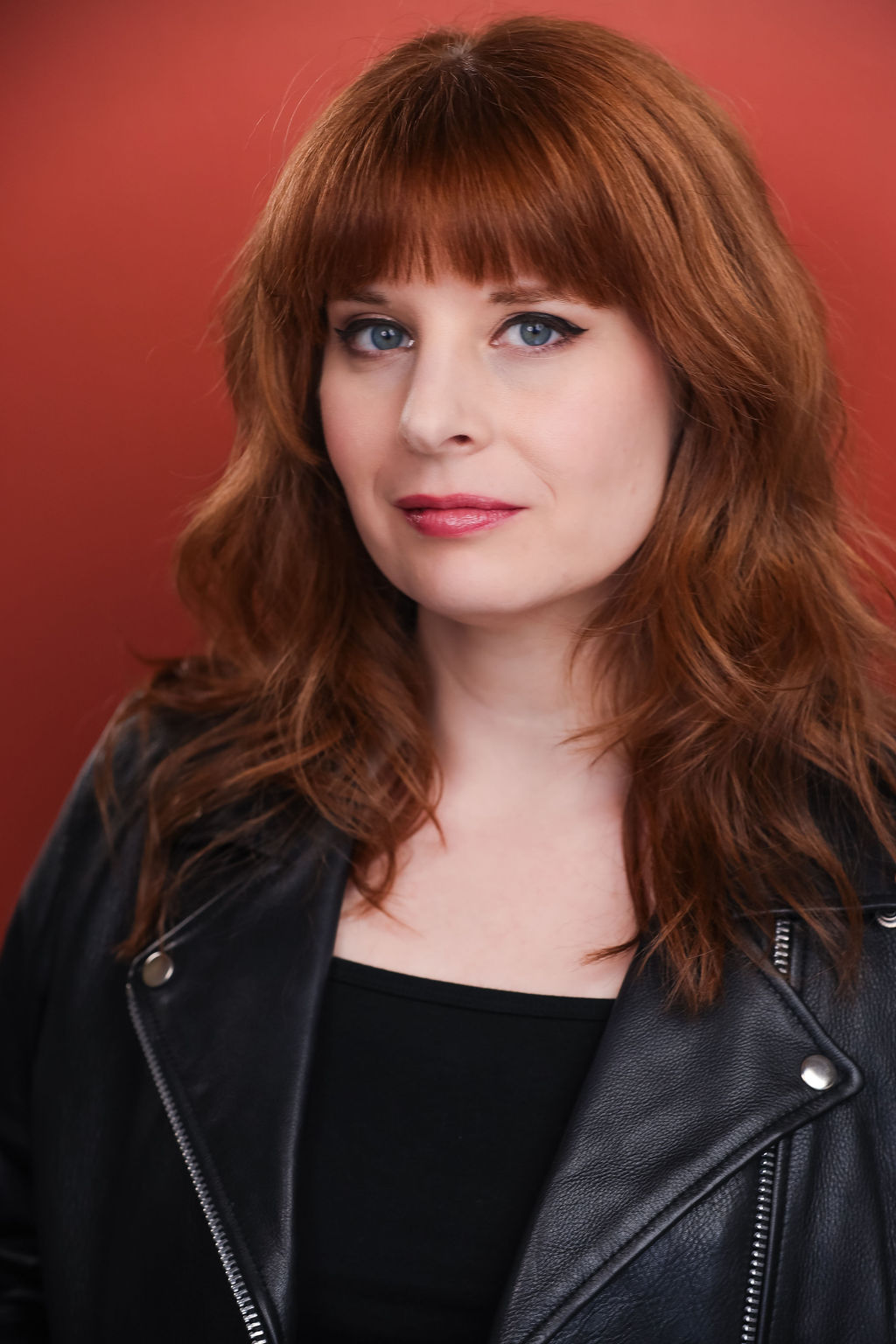 Woman with red hair and blue eyes wearing a black leather jacket against a red background.