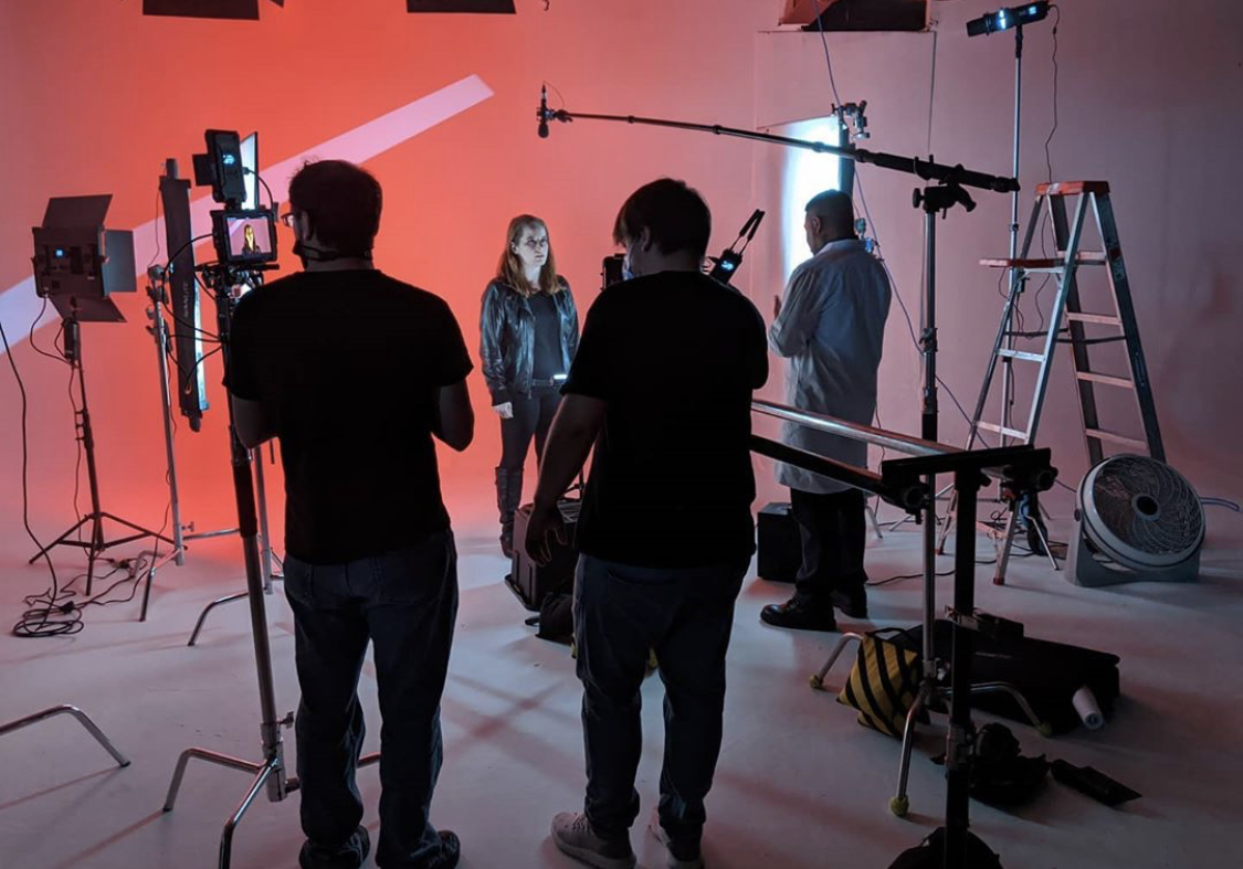 Group of people in a studio with lighting and equipment, preparing for filming or photography, with a red background.
