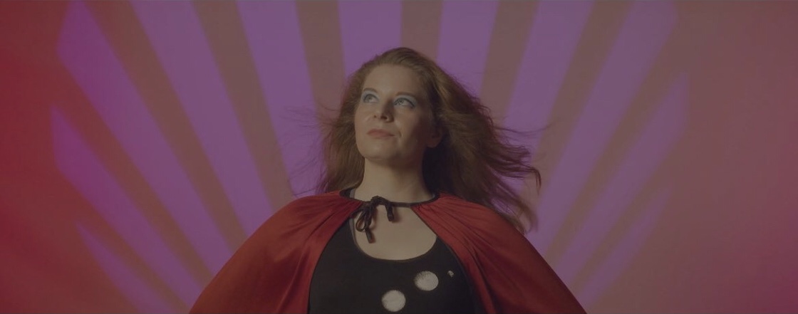 Woman with flowing hair wearing a red cape and black dress with white dots, standing against a colorful background.