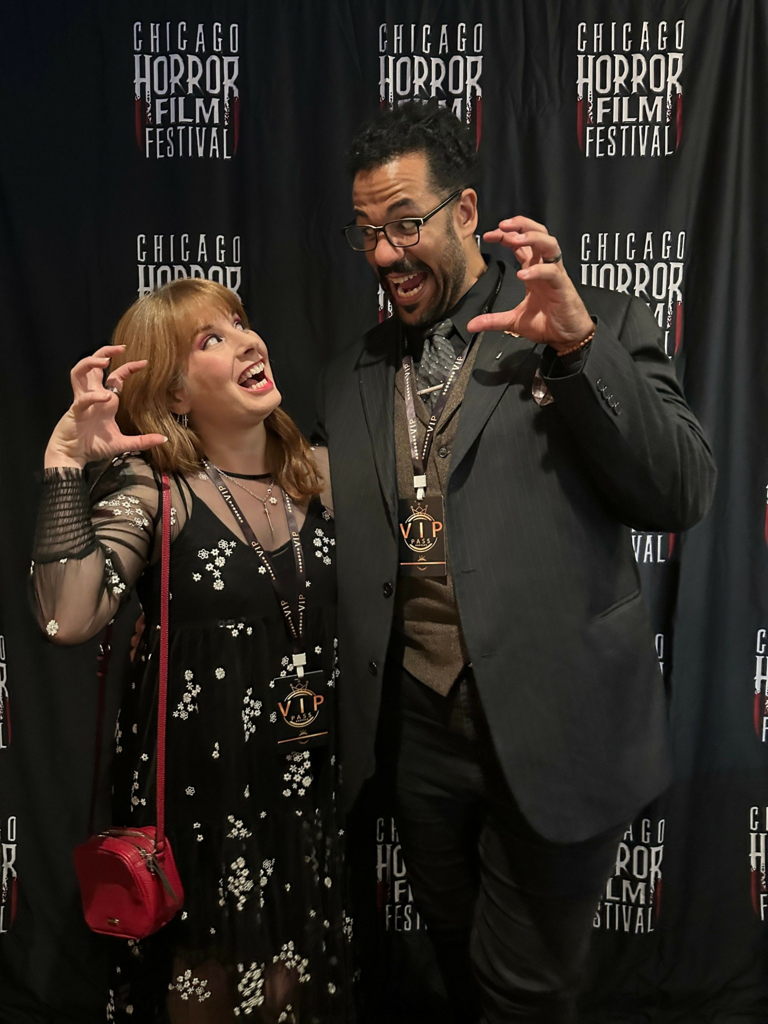 Man and woman posing together at a film festival event, with a black backdrop and text. The man is wearing glasses and a suit, the woman has blonde hair and is smiling, holding a red purse.