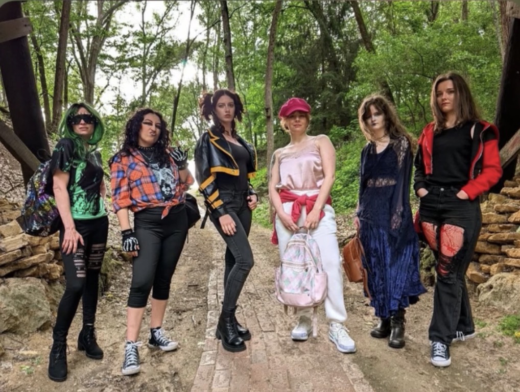 Group of six women standing outdoors on a dirt path surrounded by trees, dressed in casual clothing, some with backpacks.