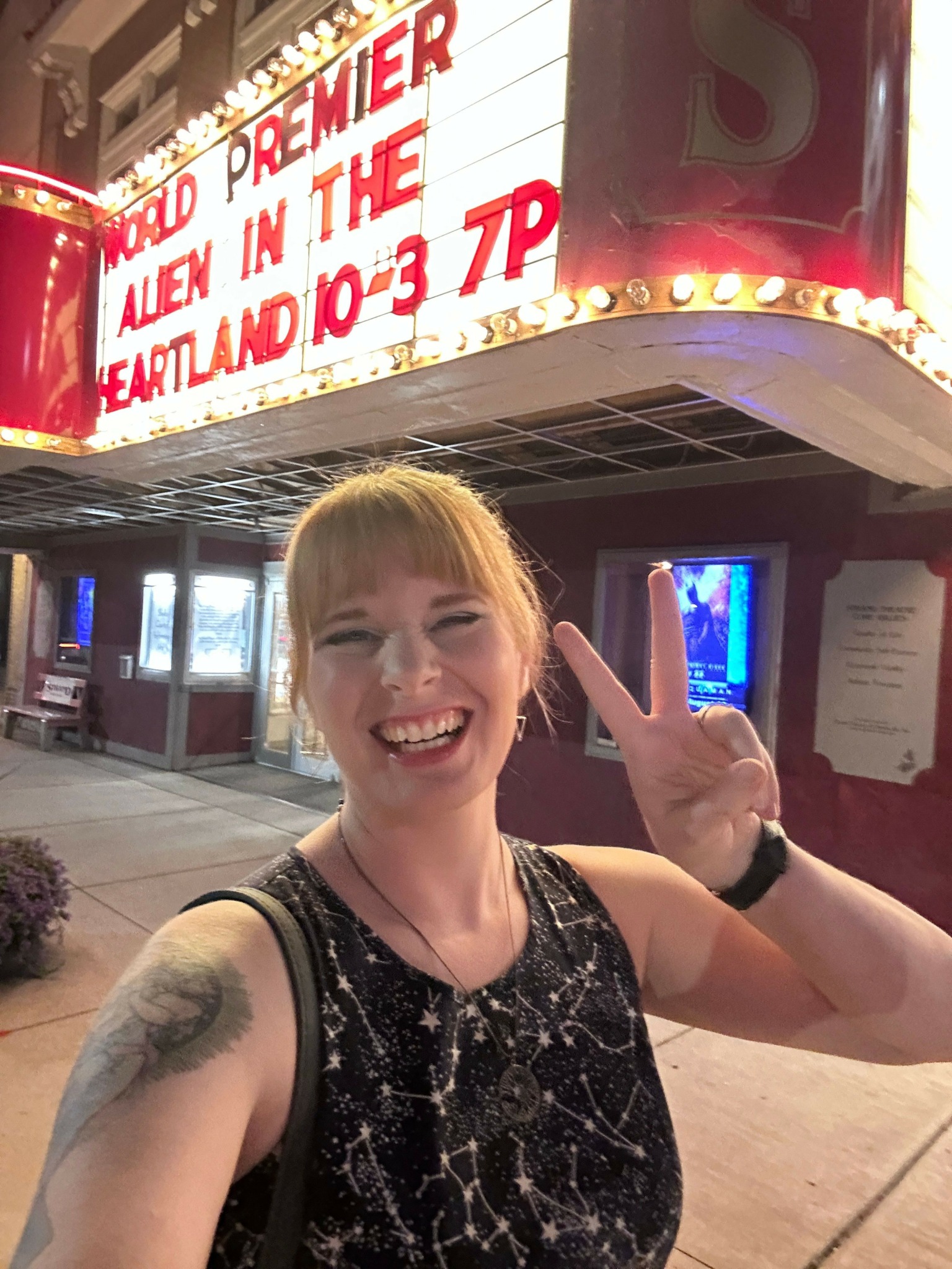 Woman with blonde hair smiling and making a peace sign, sitting indoors with a TV and a sign in the background.