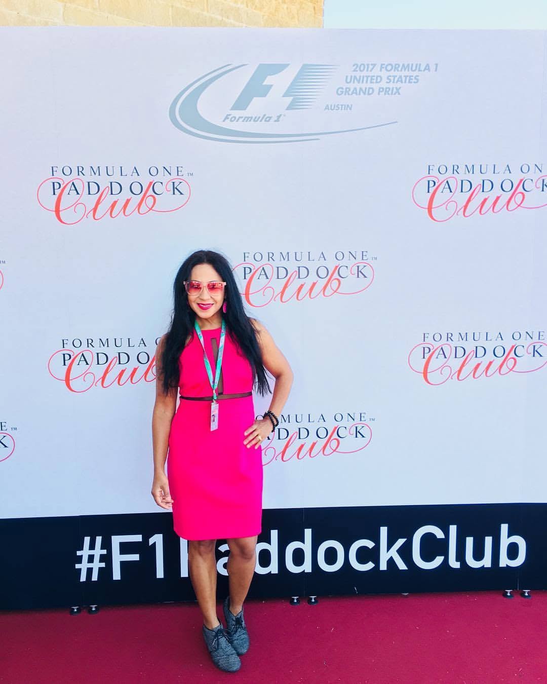 Woman in pink dress and glasses standing in front of a backdrop with Formula One and paddock club logos.