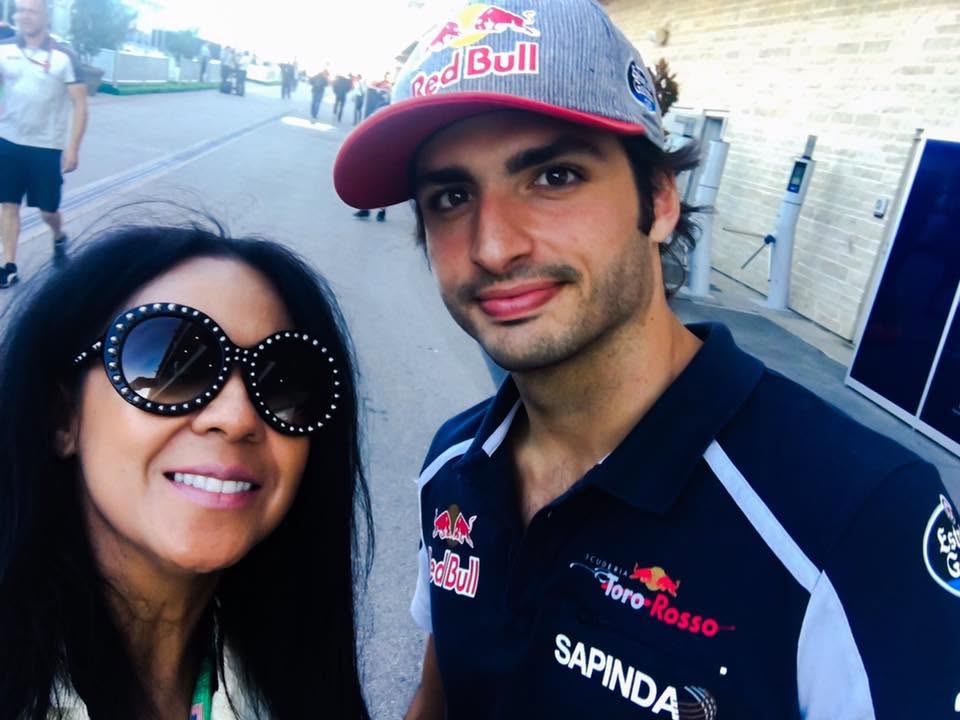 Two people smiling outdoors, one wearing large sunglasses and the other wearing a Red Bull cap and racing team shirt.