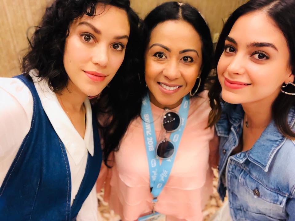 Three women smiling closely for a selfie, wearing casual clothing, with a blurred background.