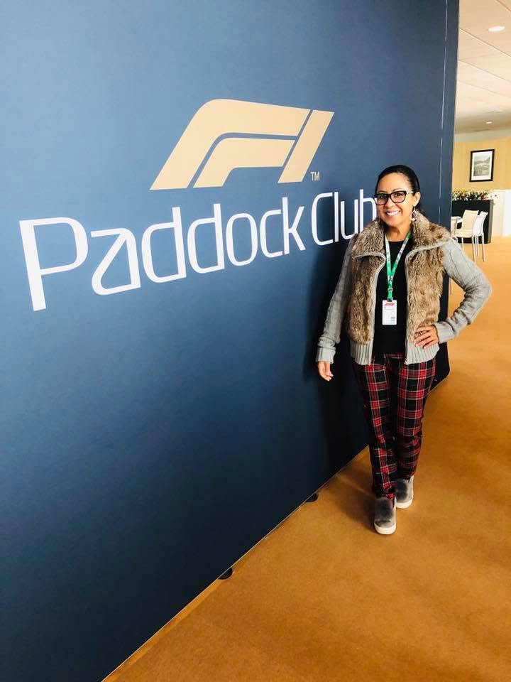 Person standing next to a blue wall with 'Paddock Club' and a logo, smiling, wearing glasses, a jacket, and plaid pants.