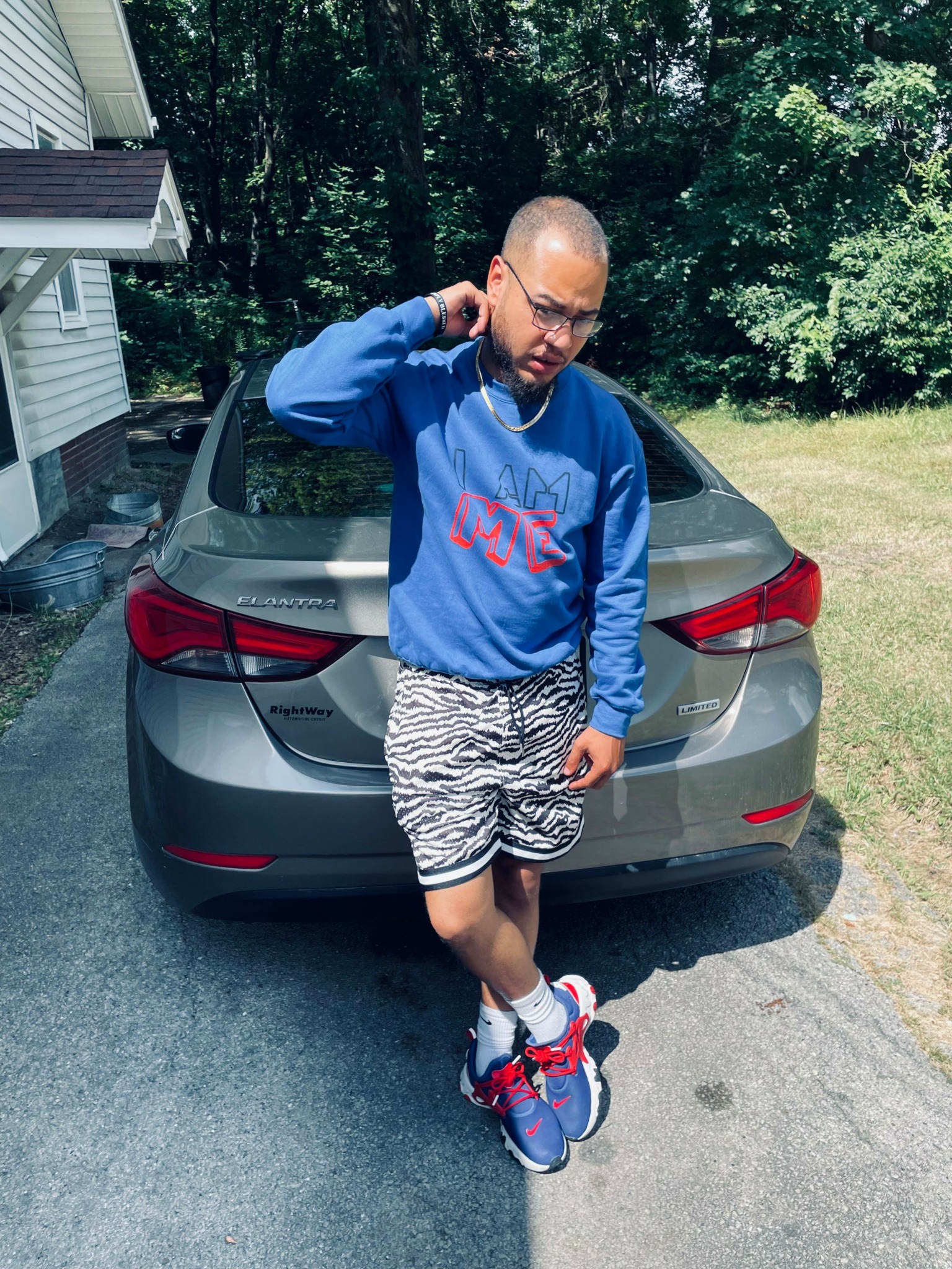 Person standing in front of a gray car, wearing a blue sweatshirt, zebra-patterned shorts, and colorful sneakers, outdoors.