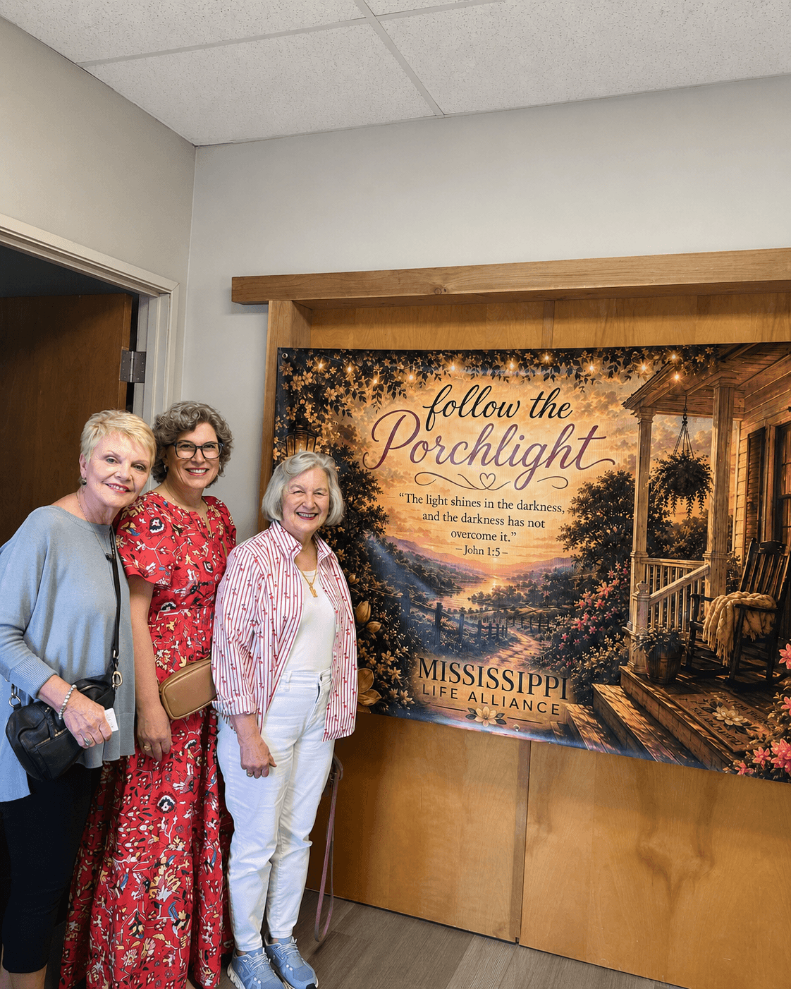 Three women standing indoors next to a large poster with a porch scene and sunset, smiling at the camera.