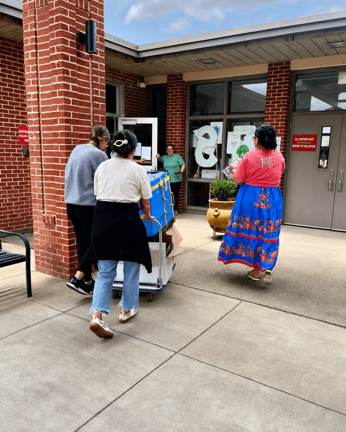 Group of people outside a brick building, some pushing a cart, others walking, with large windows and a door in the background.