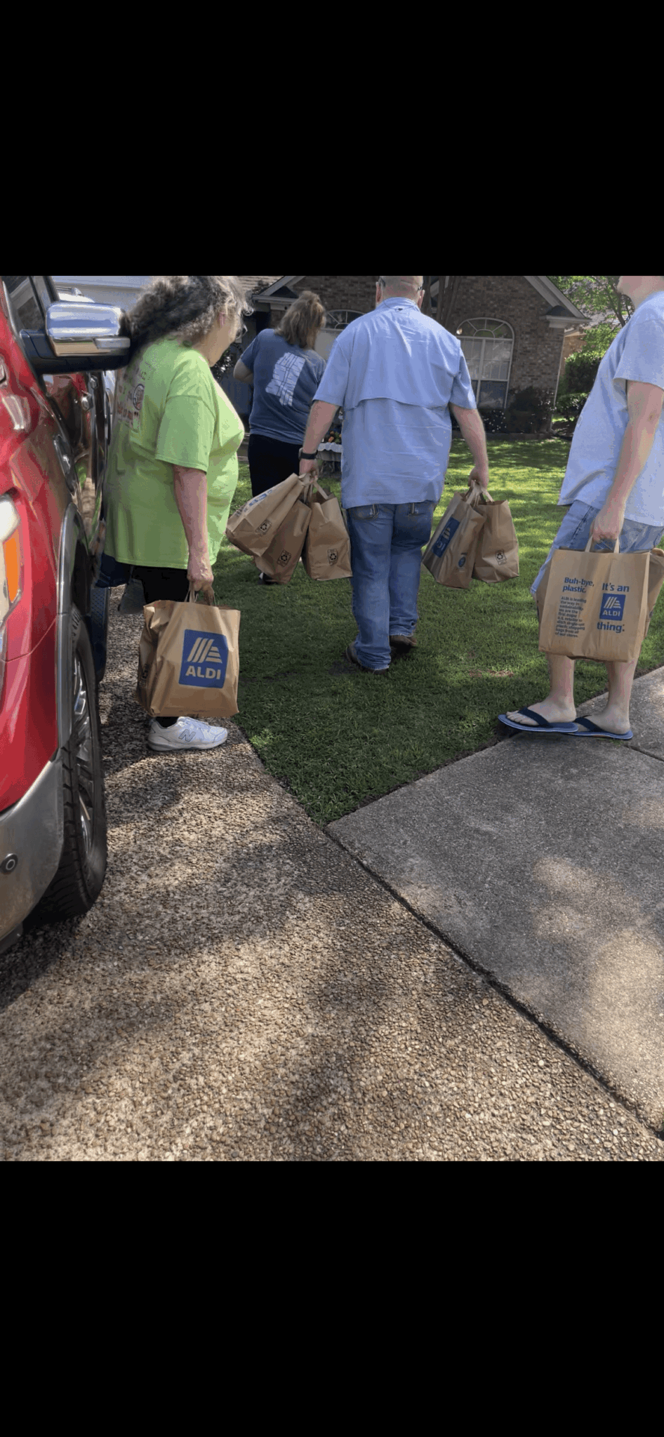 People standing outdoors holding paper bags, some wearing casual clothing, near a sidewalk and a parked vehicle.
