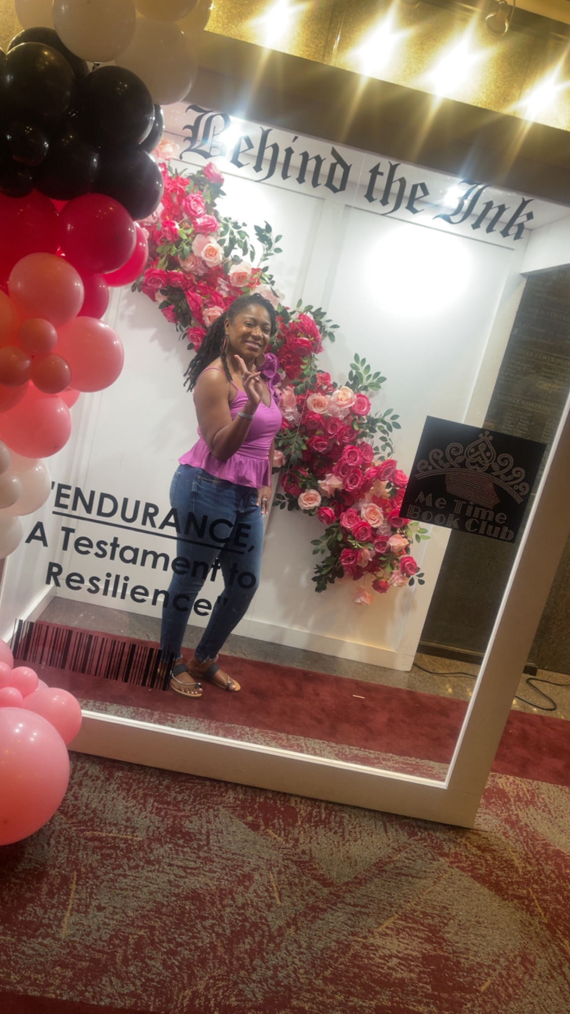Woman taking a selfie in front of a floral backdrop with pink flowers and balloons, mirror reflects part of the event space.