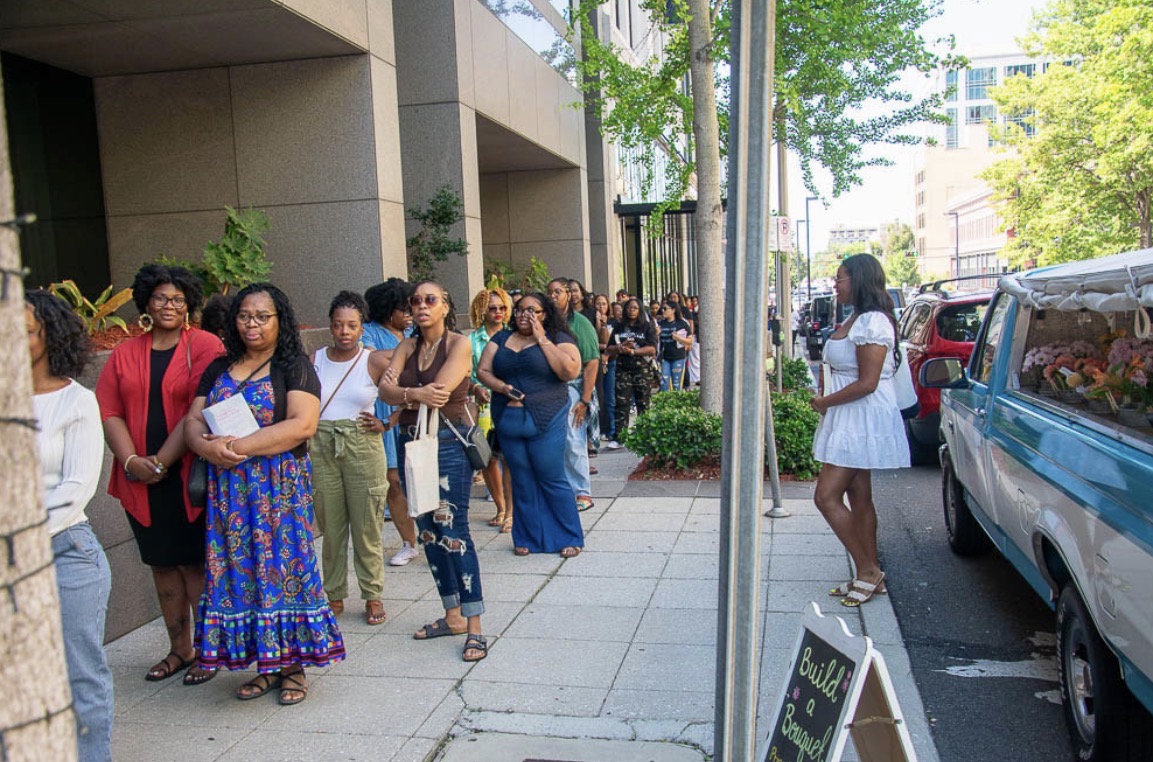 Group of people waiting in line on sidewalk outside building, woman in white dress standing near parked vehicle, trees and buildings in background.