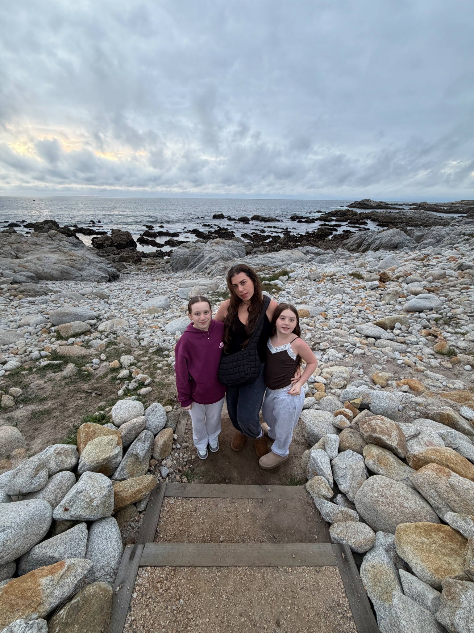 Three people standing on a rocky beach with ocean and cloudy sky in background.