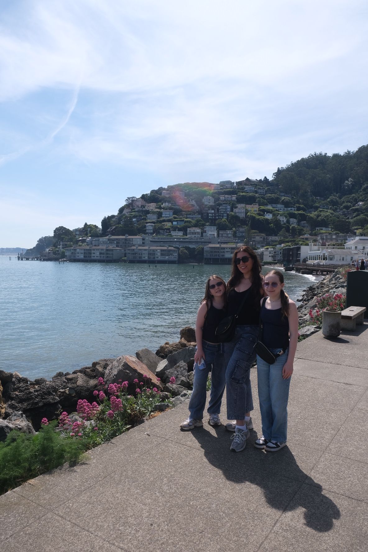Three people stand on a paved path near water with a cityscape and bridge in the background.
