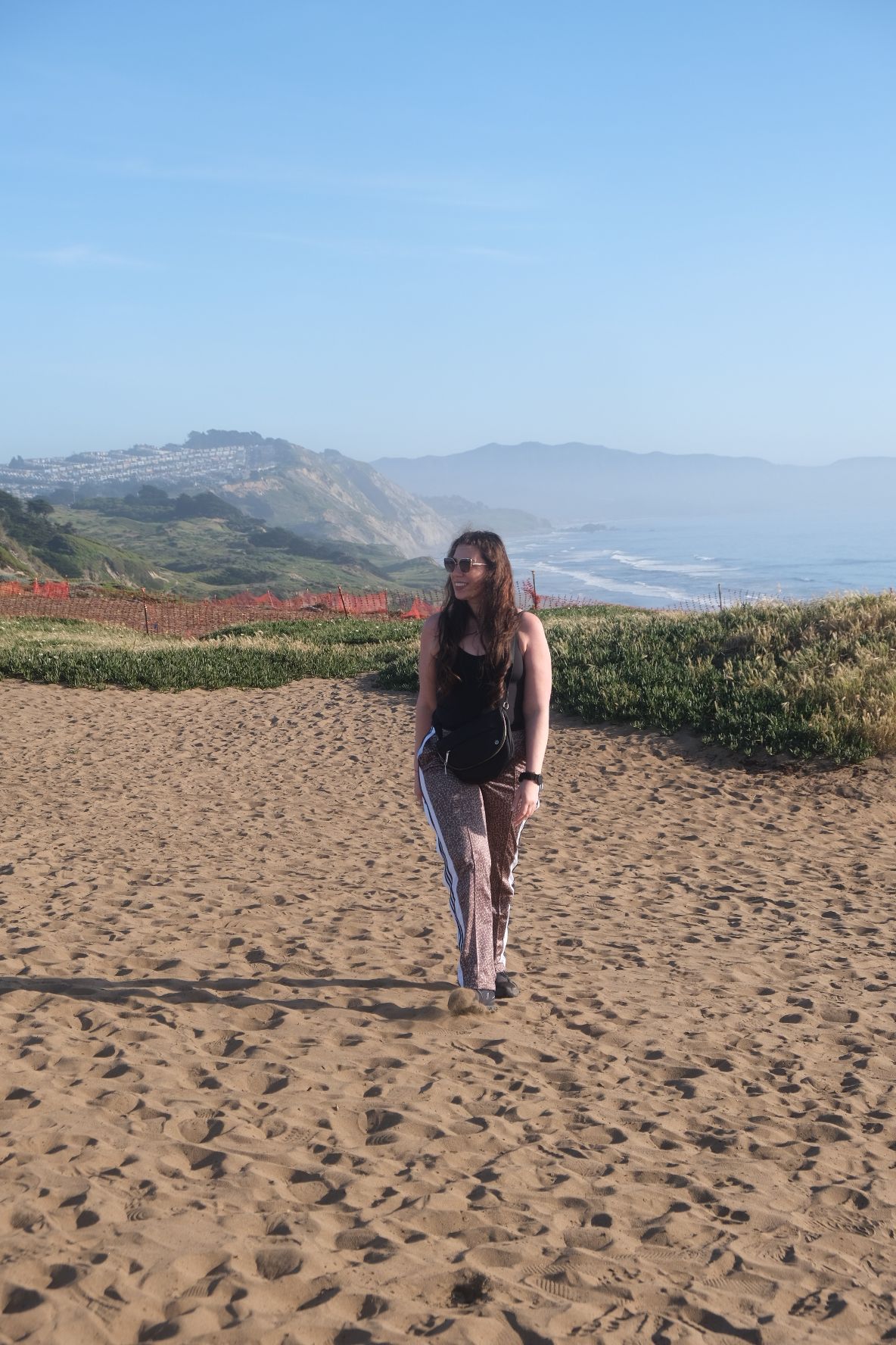 Person standing on sandy beach with mountains and blue sky in background.