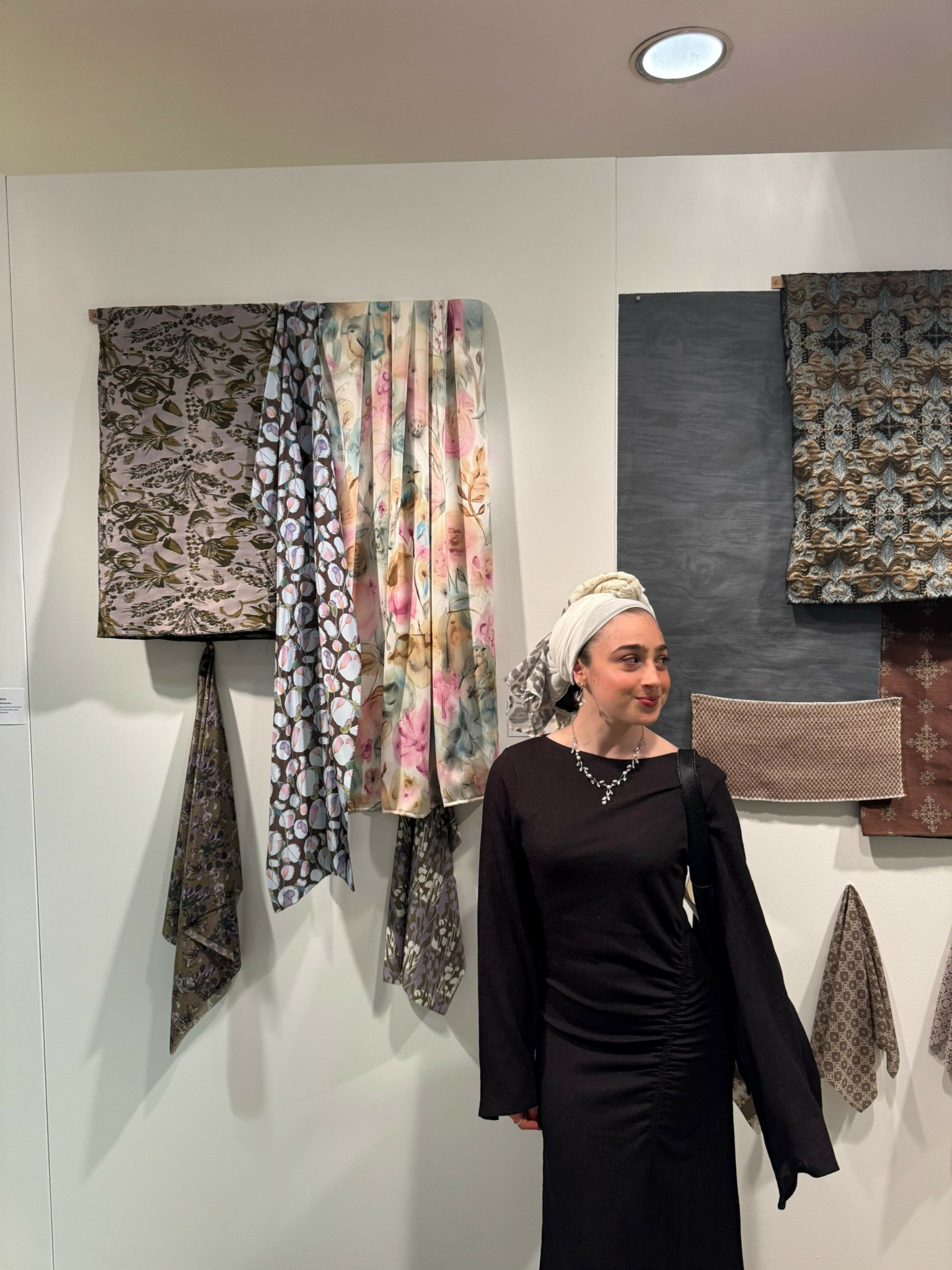 Woman in black dress and headscarf standing in front of hanging fabric samples on wall.