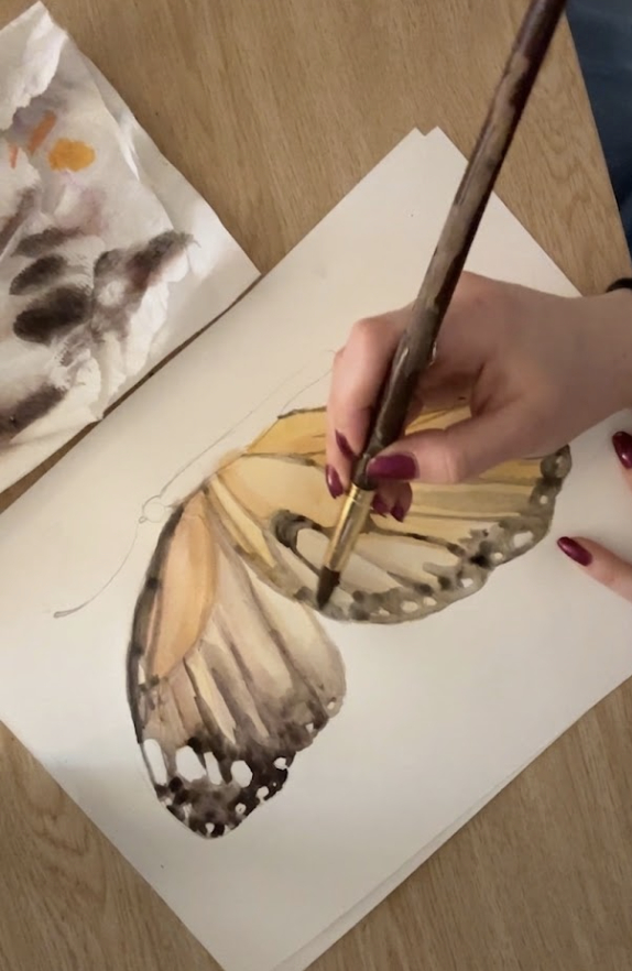 Person painting a butterfly with yellow and brown wings on paper, holding a brush, with a palette nearby.