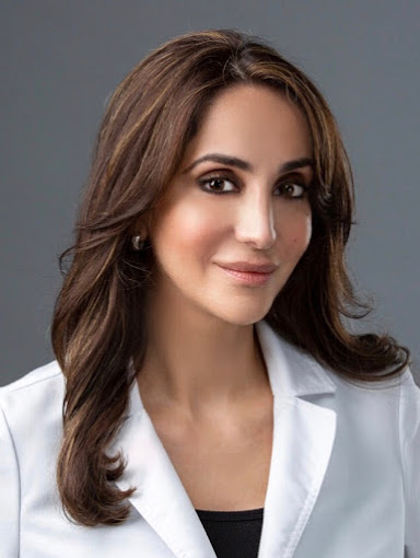 Portrait of a woman with long brown hair, wearing a white blazer, smiling softly against a gray background.