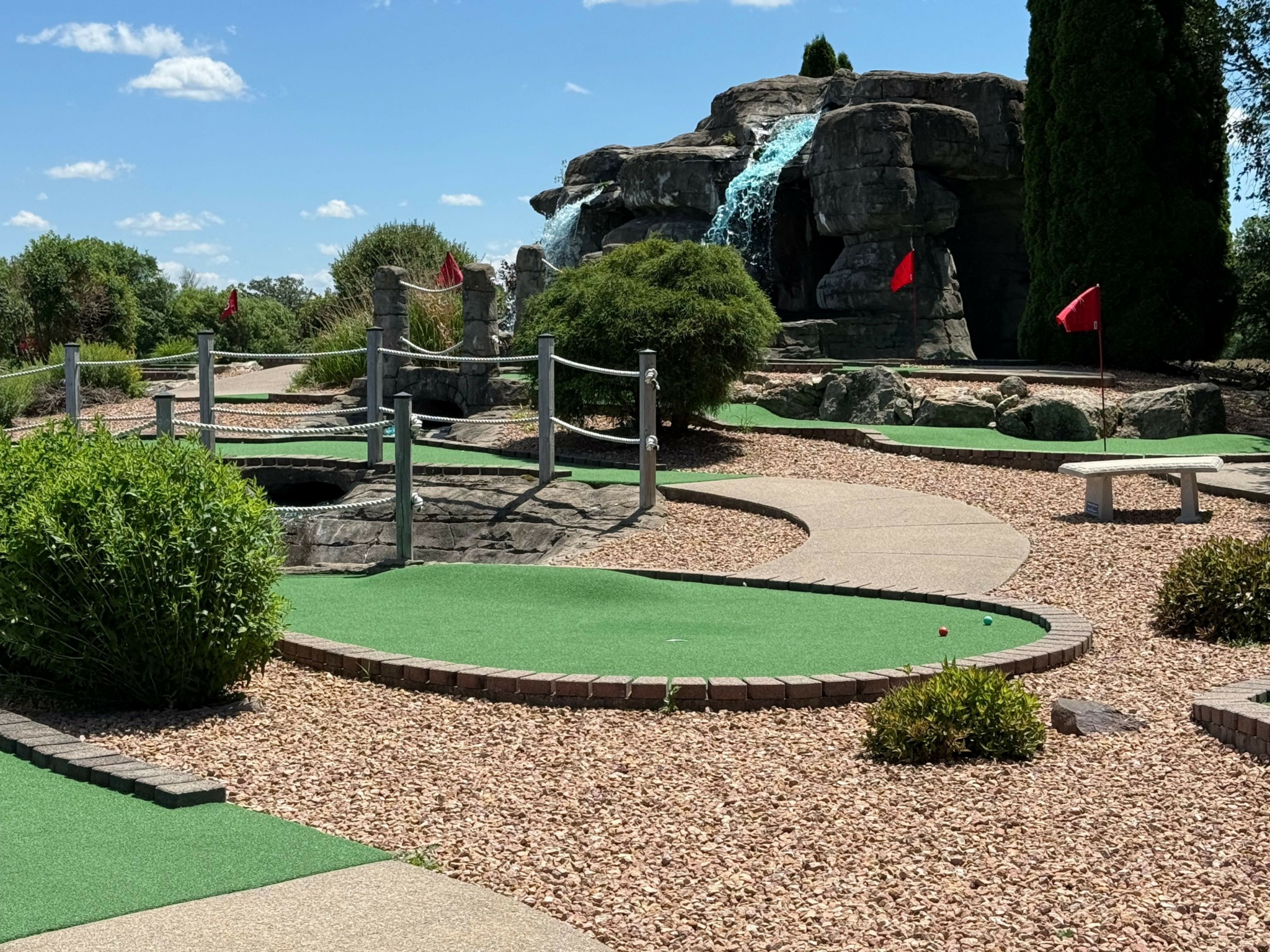 Miniature golf course with green putting areas, pathways, trees, and a small bridge, under a partly cloudy sky.