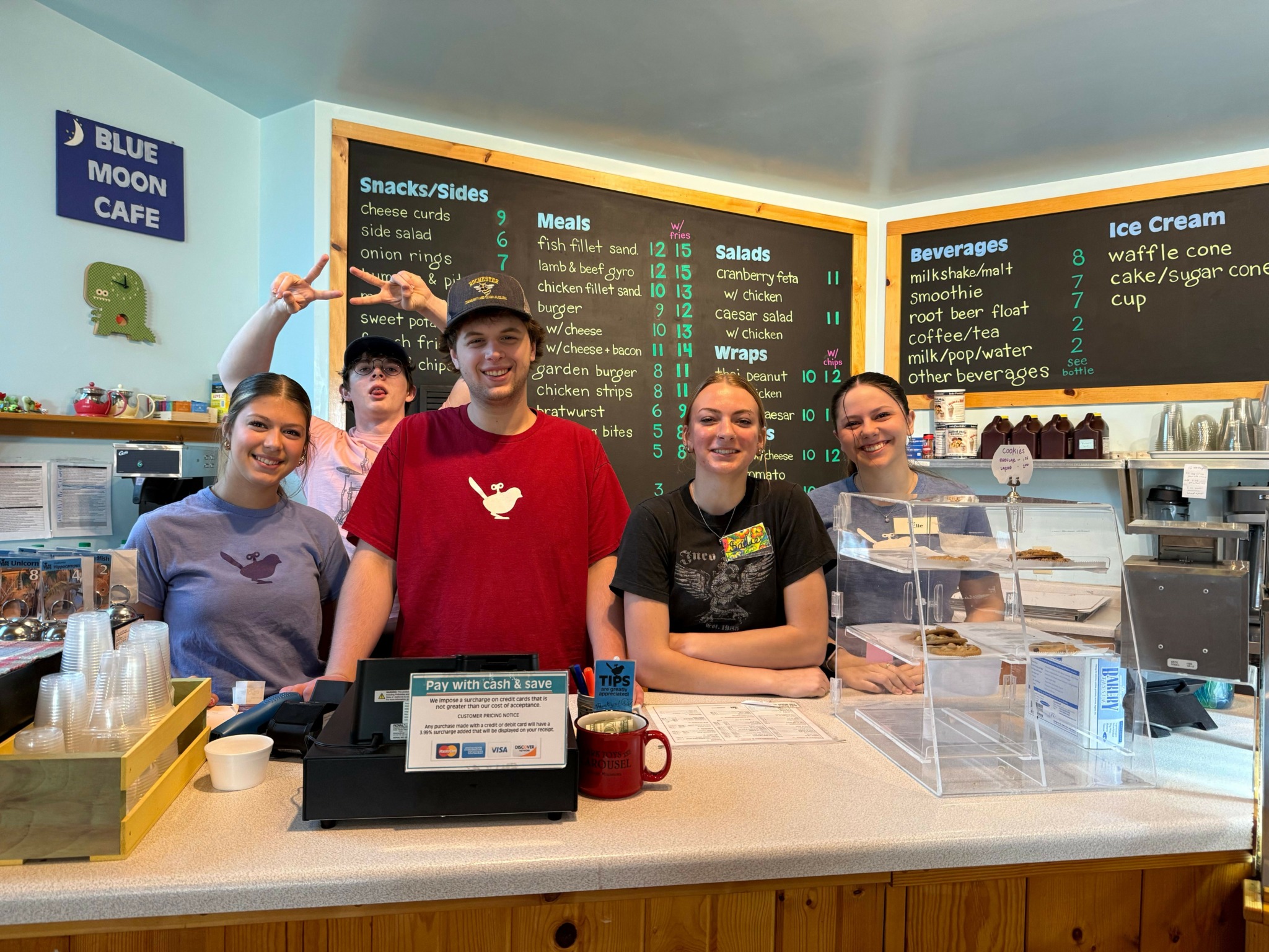 Four people standing behind a counter in a store, with a menu board and shelves in the background.