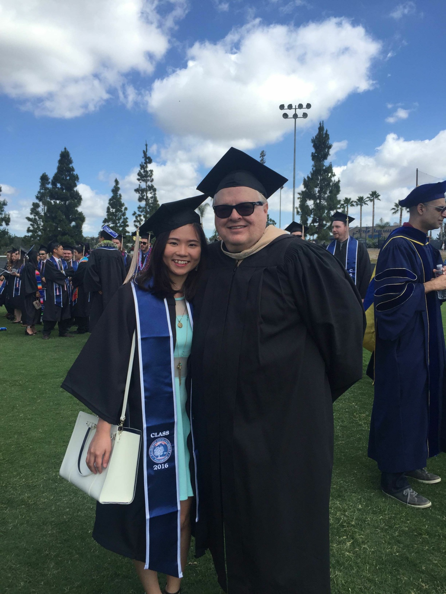 Two graduates in caps and gowns smiling outdoors, with a crowd and trees in the background.