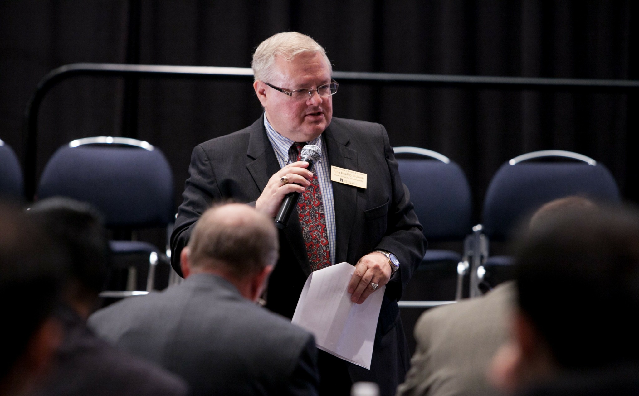 Elderly man in suit speaking into a microphone at a formal event, holding papers, with chairs and audience in background.