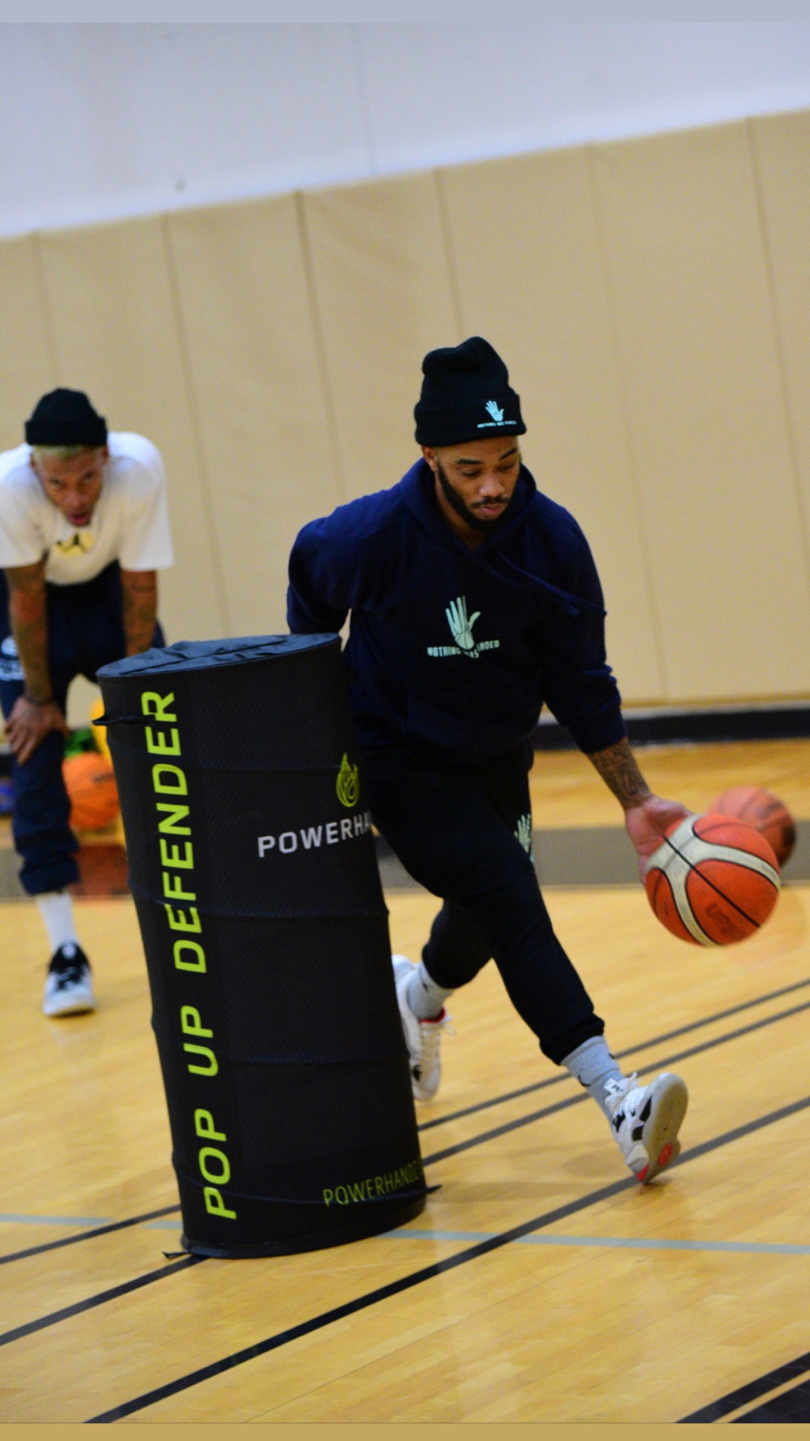 Person in dark hoodie dribbling basketball around a padded obstacle on a gym floor, with another person in the background.