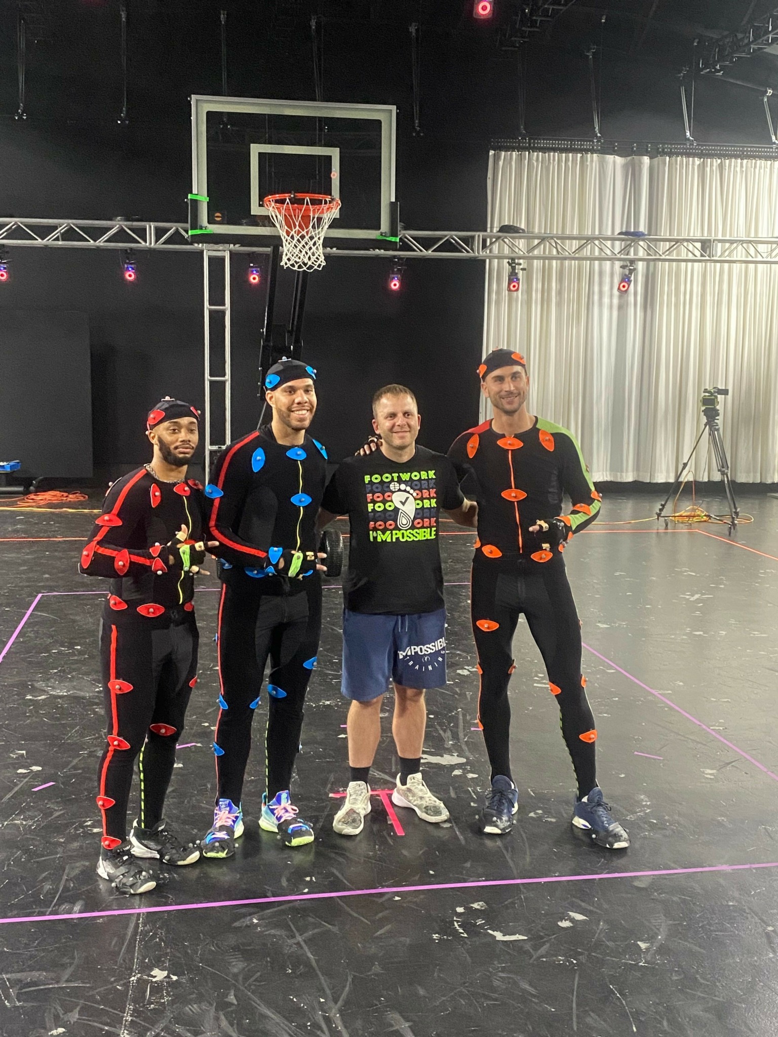 Four people stand on a basketball court near a hoop, smiling, with one person holding a basketball. Indoor setting with curtains in background.