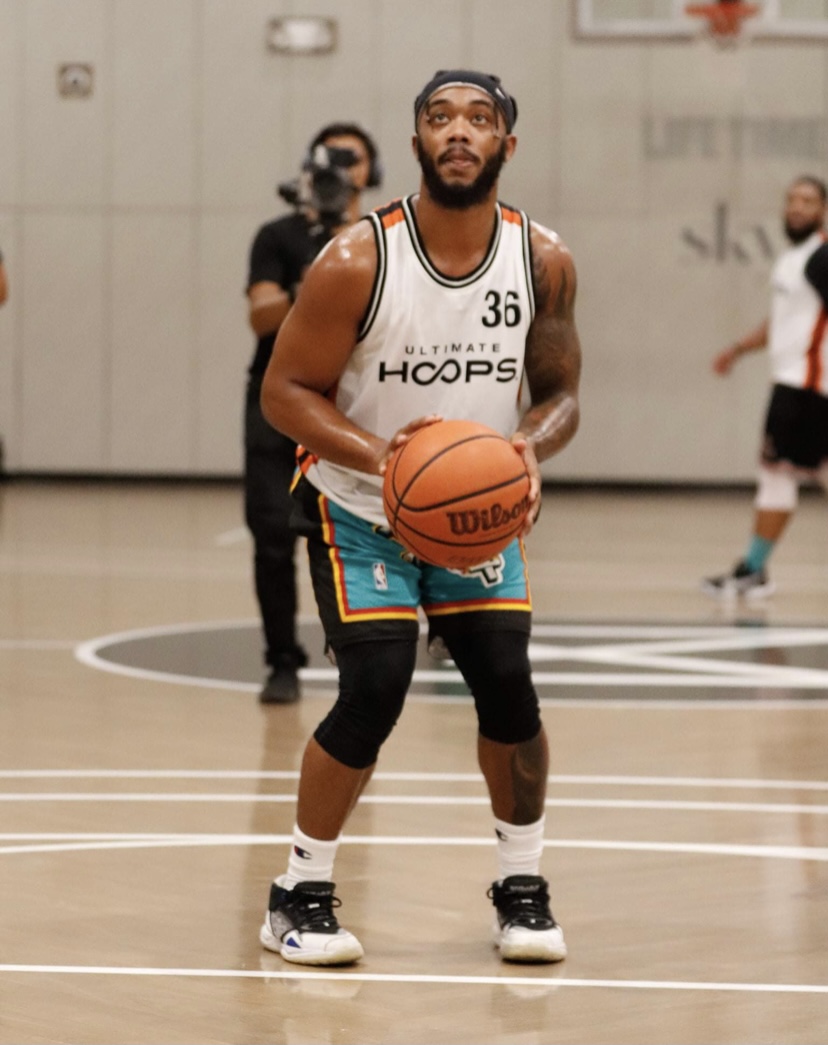 Basketball player in a white jersey holding a basketball on court, with teammates and staff in background.